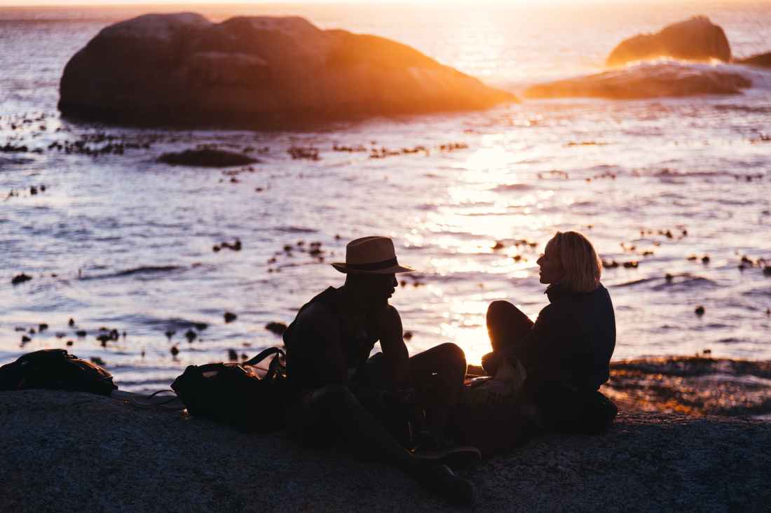 man and woman sitting on shore near body of water during sunset